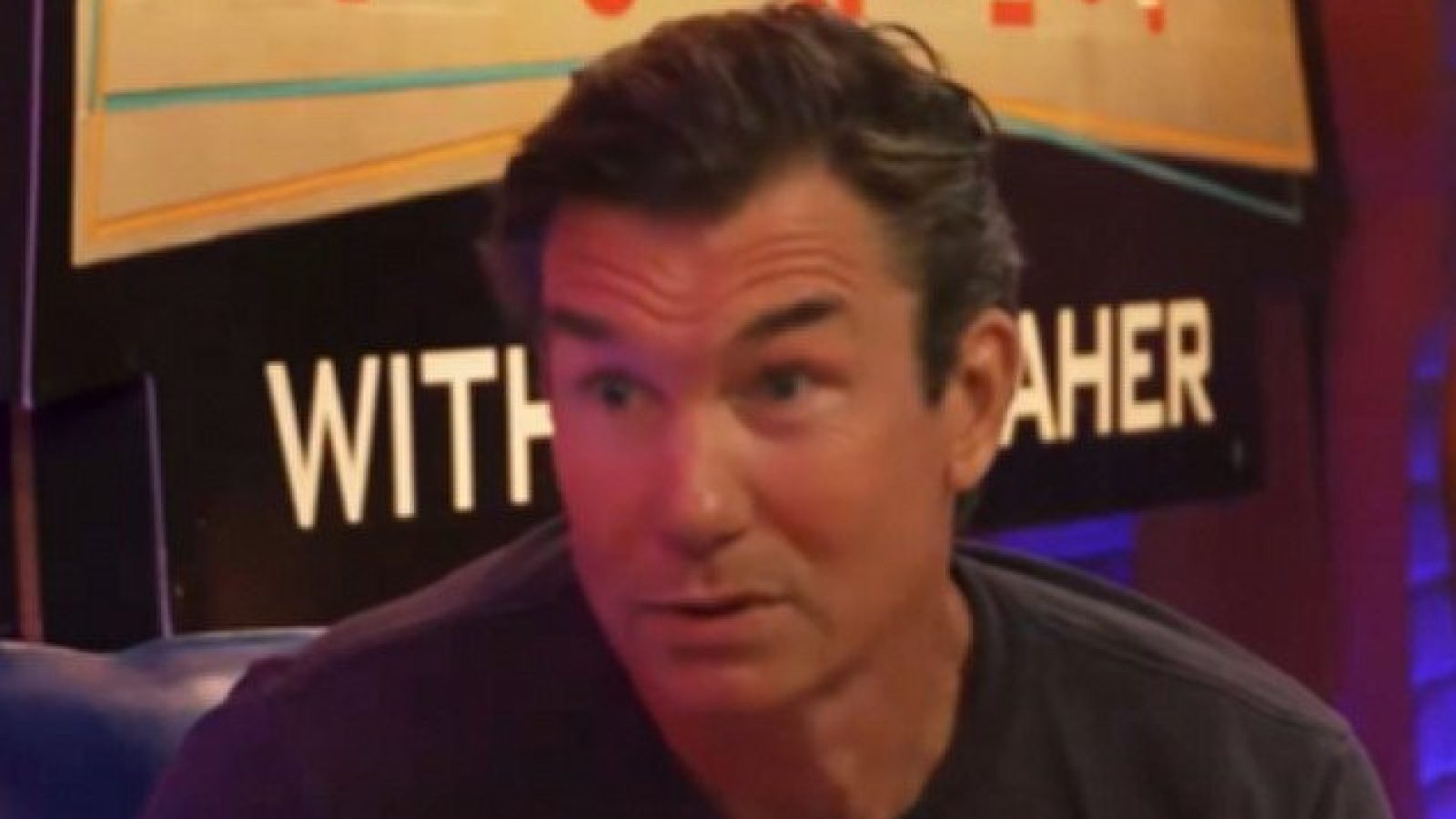 Man with dark hair speaking animatedly during a talk show, with a colorful backdrop featuring the show's title.