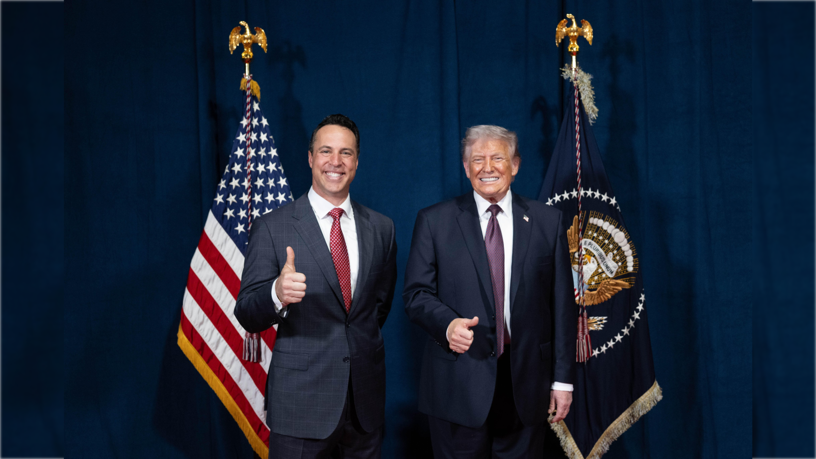 Two men smiling and giving thumbs up in front of American flags and a presidential seal backdrop.