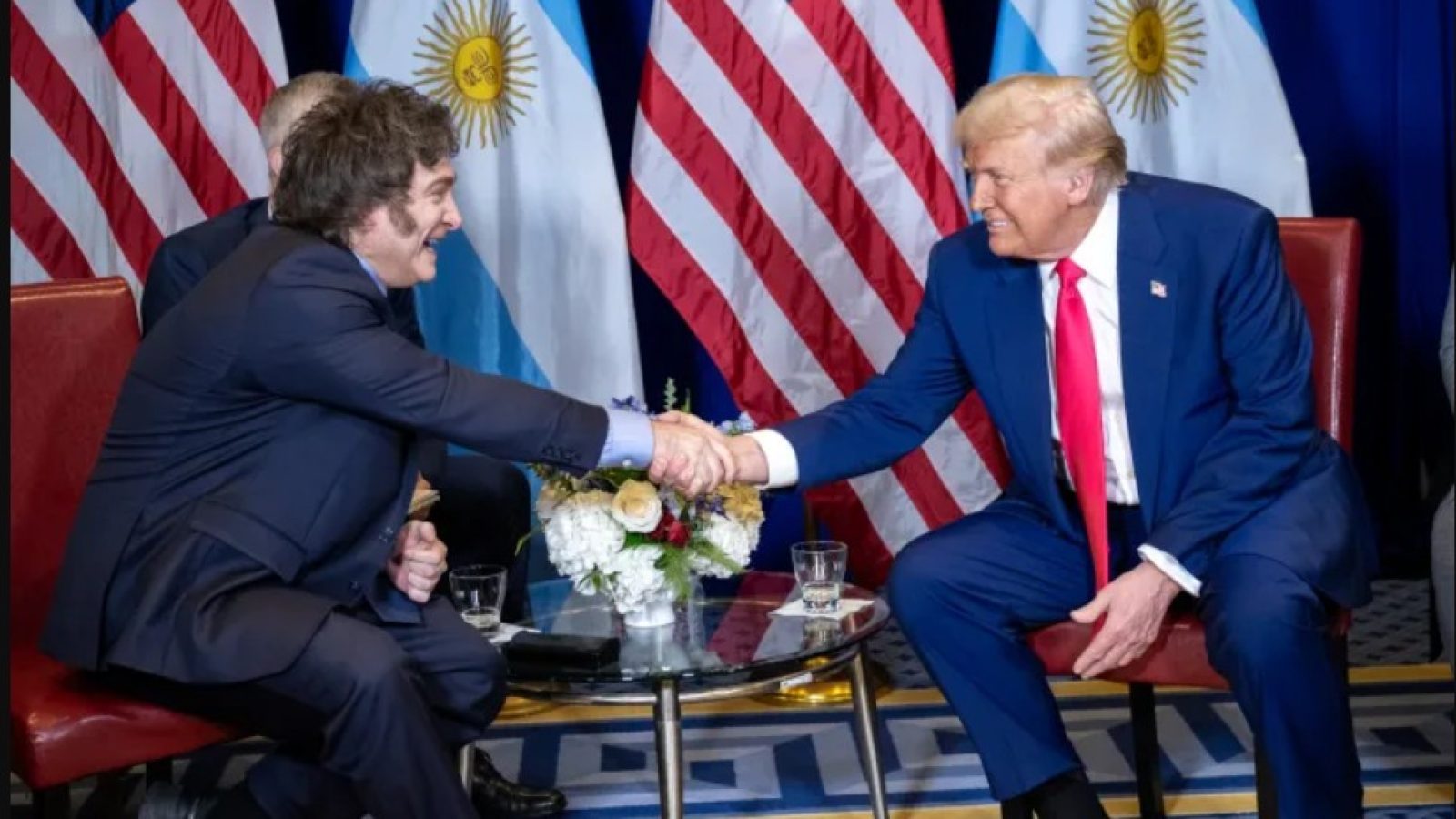 Trump and Argentine leader shake hands during a diplomatic meeting, with flags of Argentina and the USA in the background.