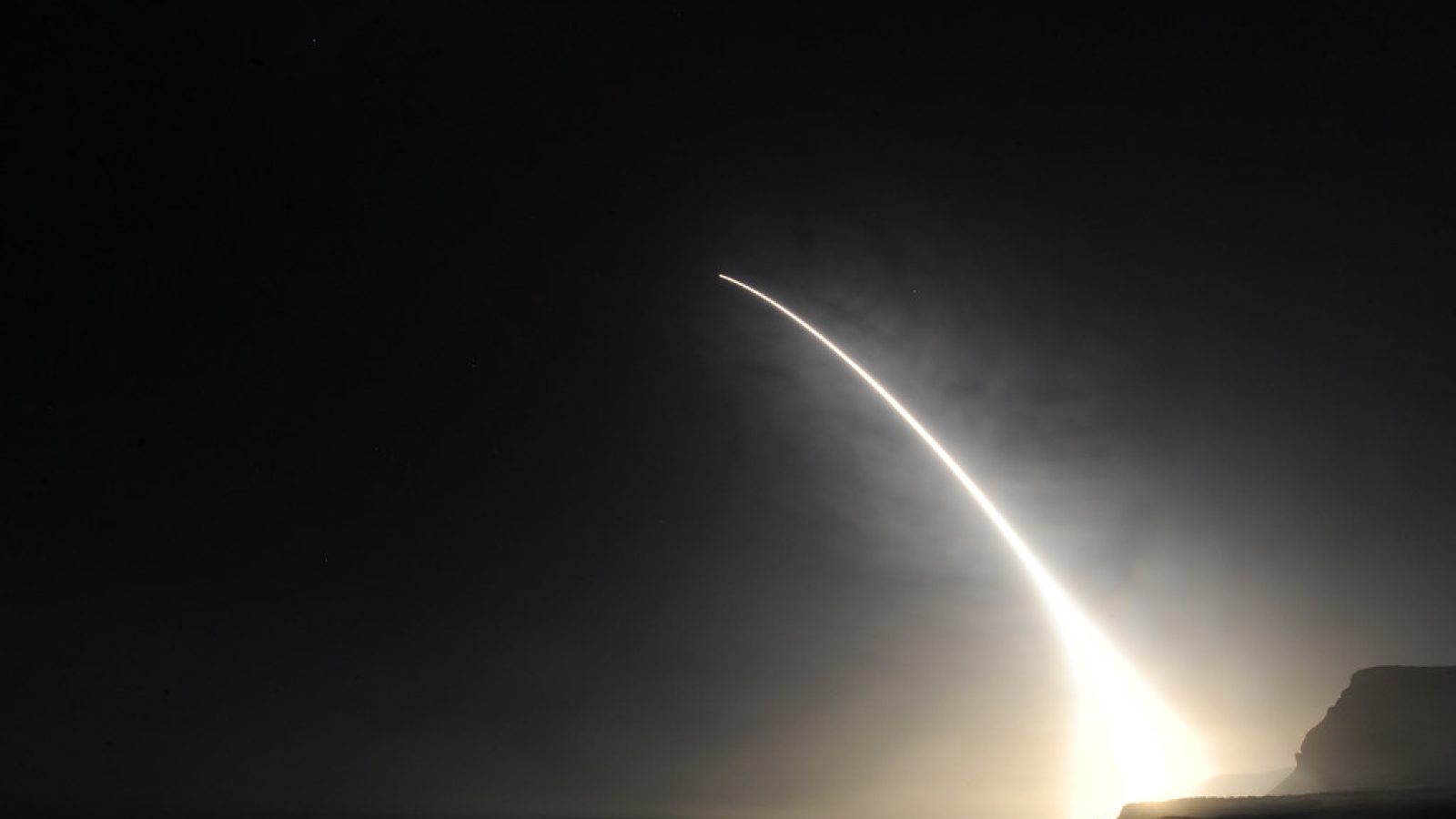 Rocket launch illuminating the night sky over a coastal landscape, showcasing the trajectory of the ascending missile against a dark backdrop.