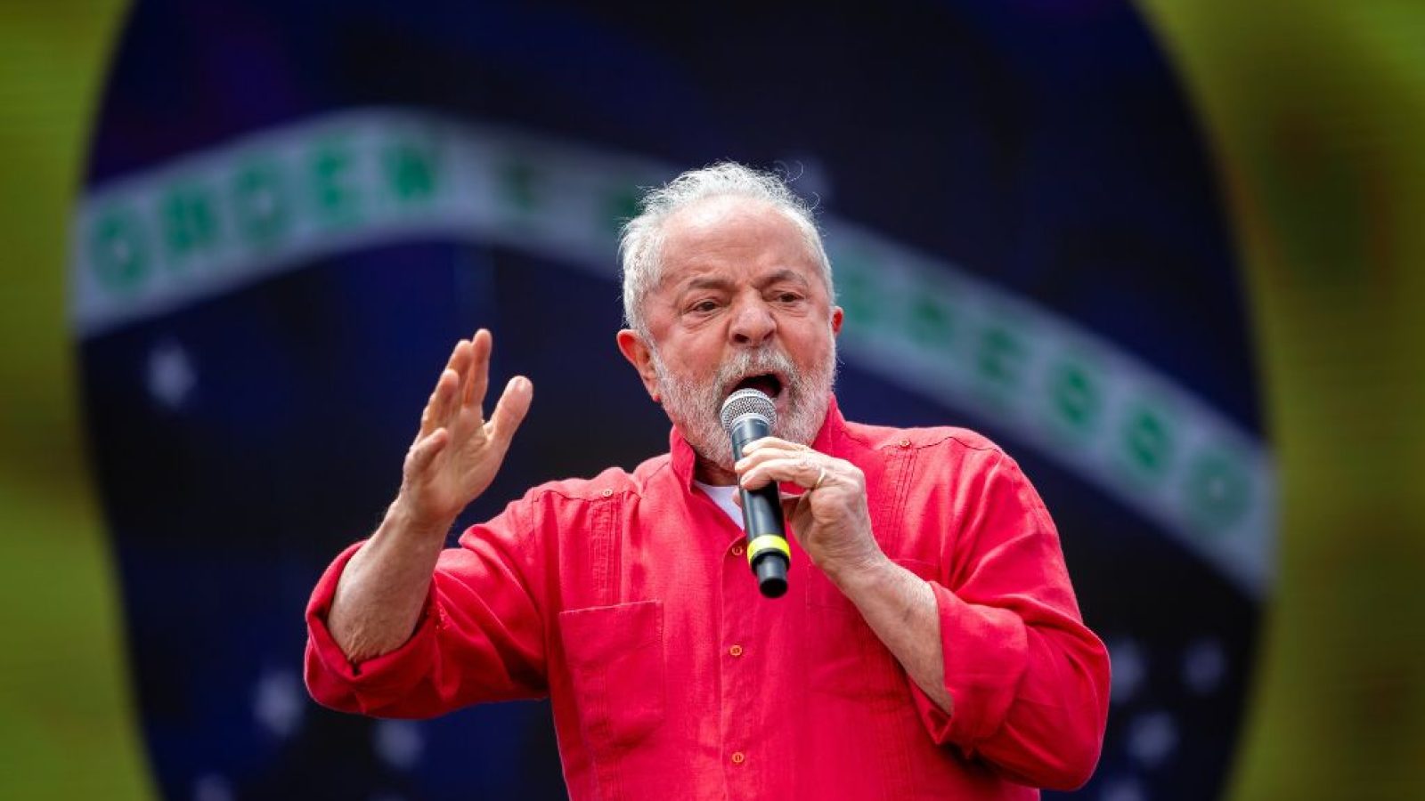 A man in a red shirt passionately speaks into a microphone at a public event with a colorful background.