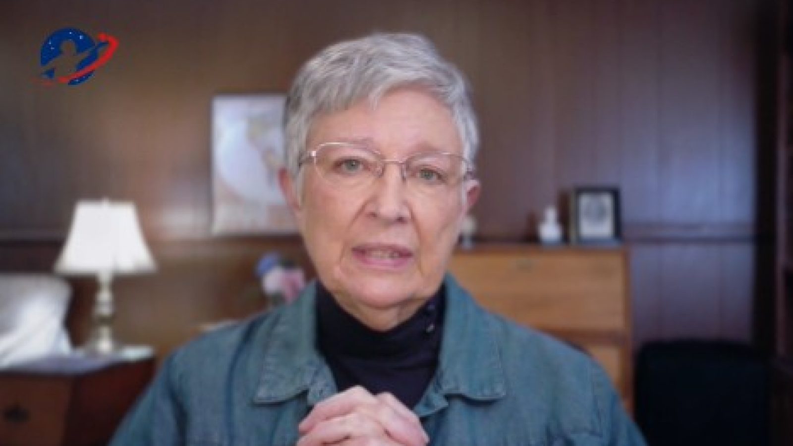 Elderly woman with short gray hair and glasses speaking in a cozy room, conveying a message of importance and sincerity.