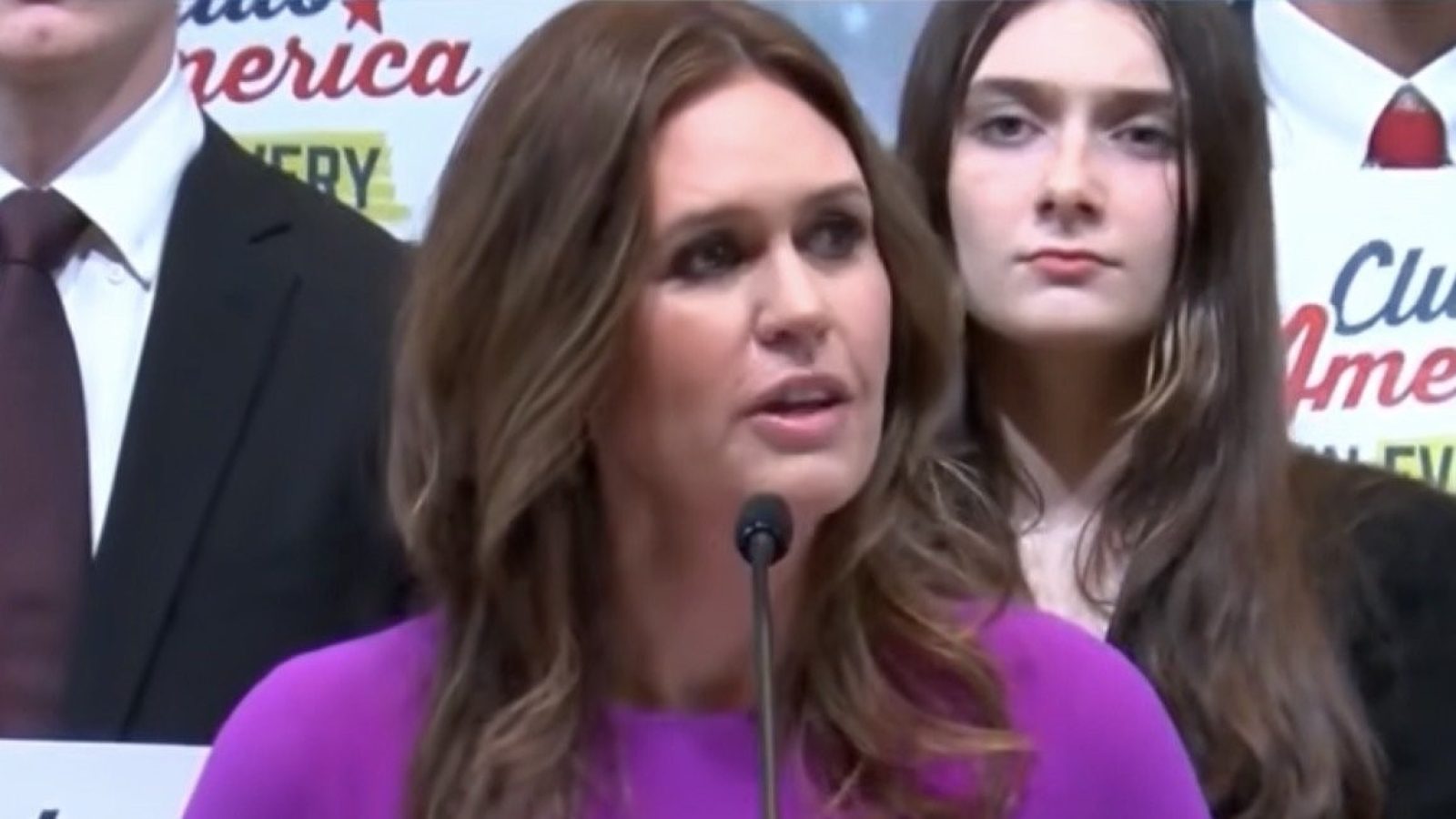 Woman in a purple dress speaks at a podium, flanked by supporters holding signs, during a public event.