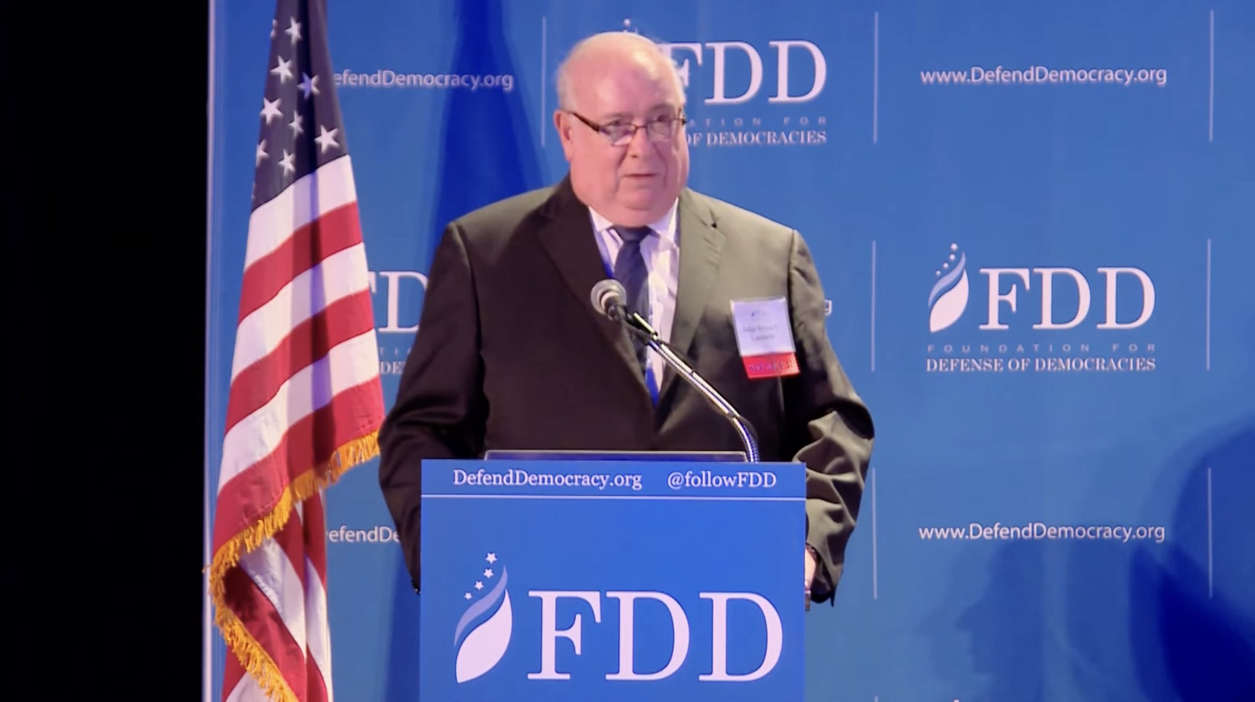 Speaker addressing the audience at the Foundation for Defense of Democracies event, with an American flag in the background.
