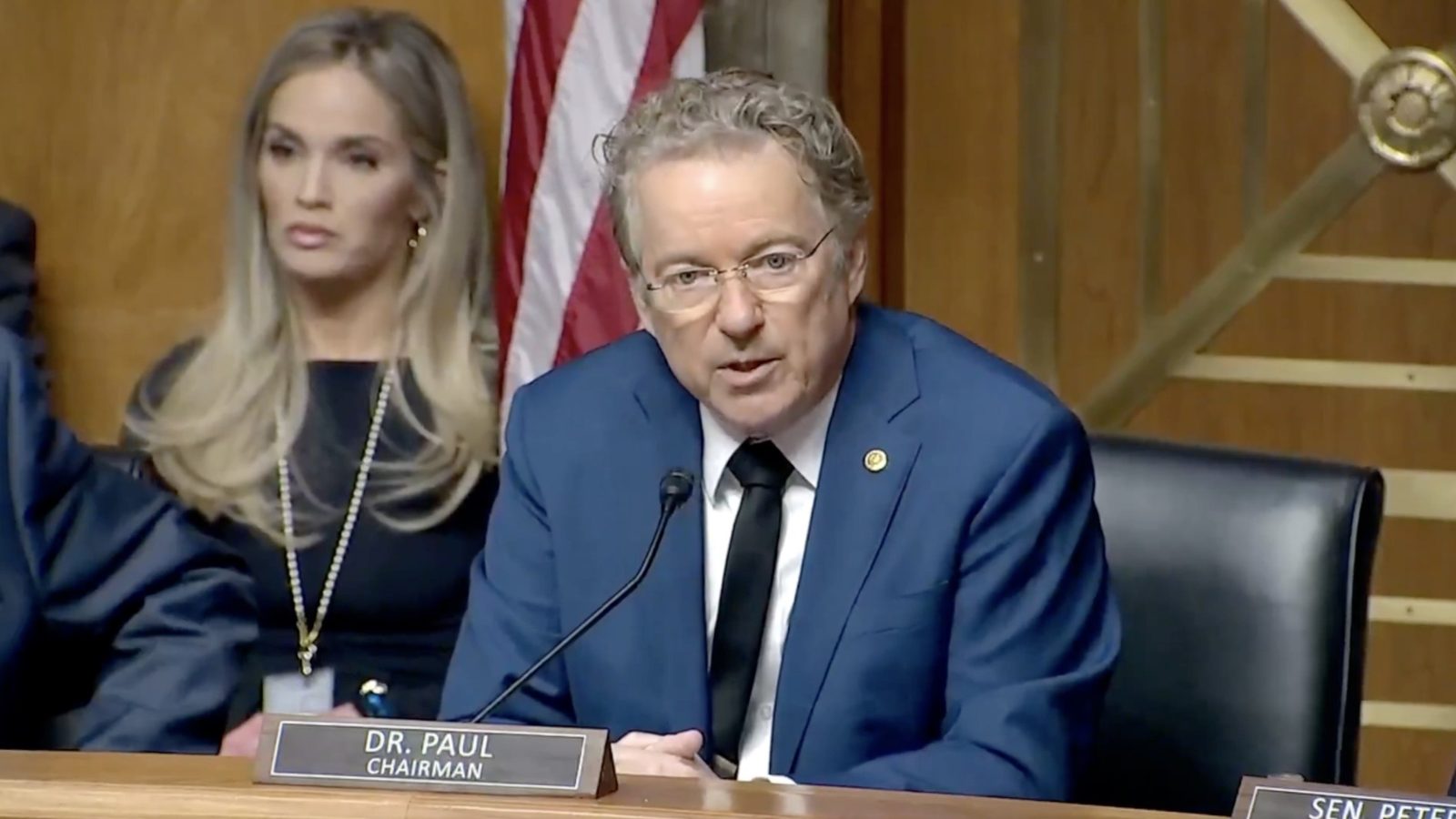 Rand Paul, Chairman, speaks at a Senate hearing with a woman in the background, showcasing a formal legislative setting.