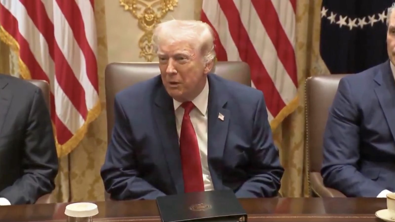 Donald Trump seated at a meeting with American flags in the background, discussing key issues with advisors.