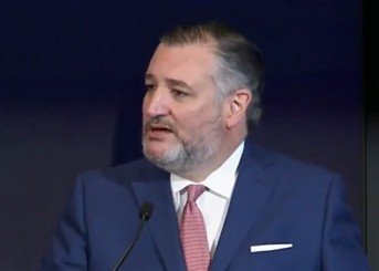Ted Cruz delivering a speech at a podium, wearing a blue suit and pink tie, addressing an audience with a serious expression.