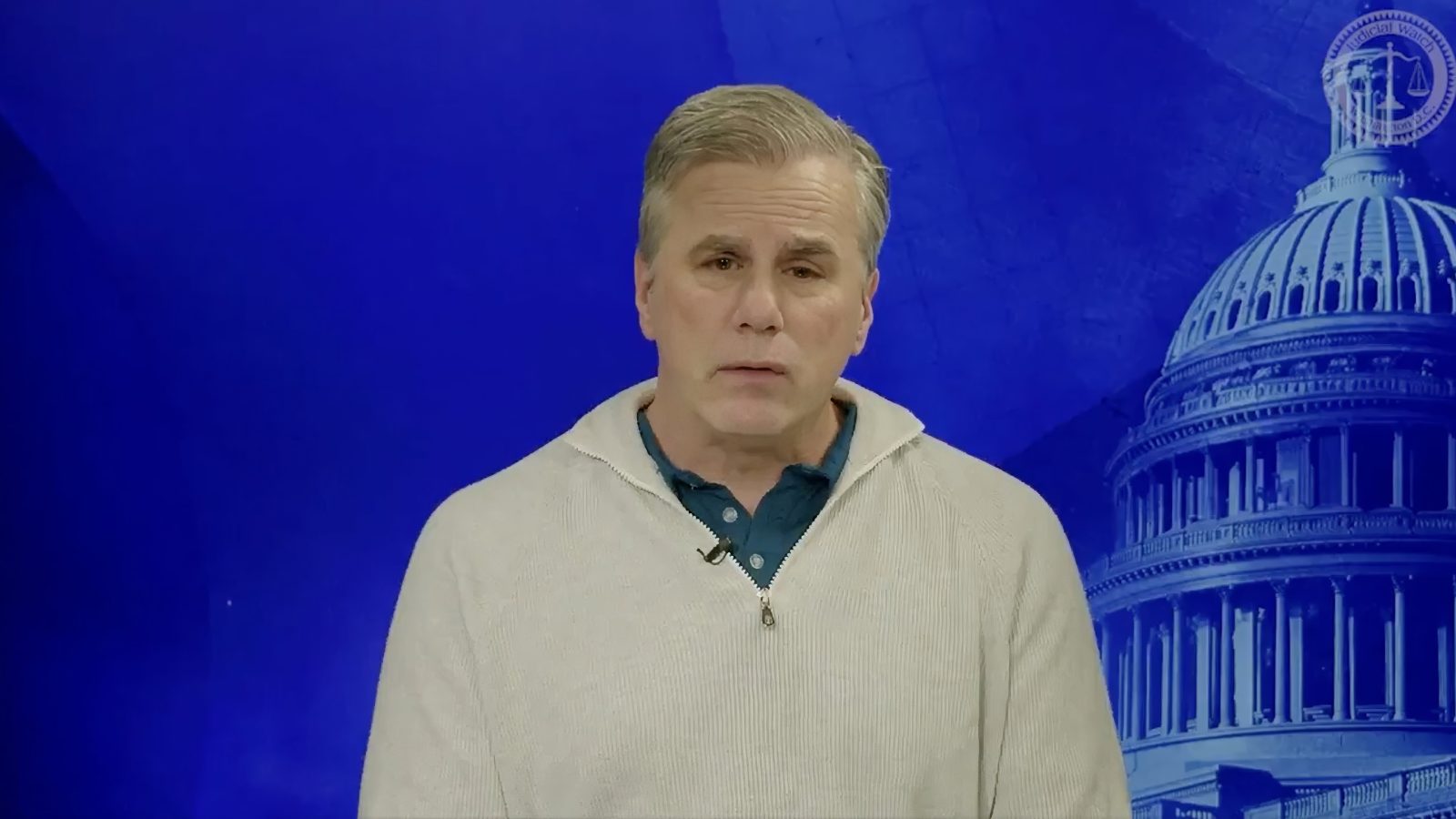 Man in a light sweater speaks against a blue background featuring the U.S. Capitol dome, discussing political issues or legal matters.
