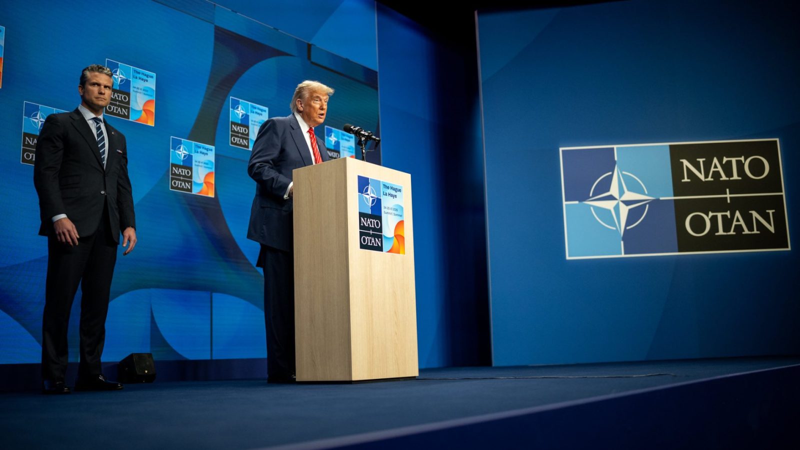 Former President Donald Trump delivers a speech at a NATO event, accompanied by an official, with the NATO logo displayed prominently in the background.