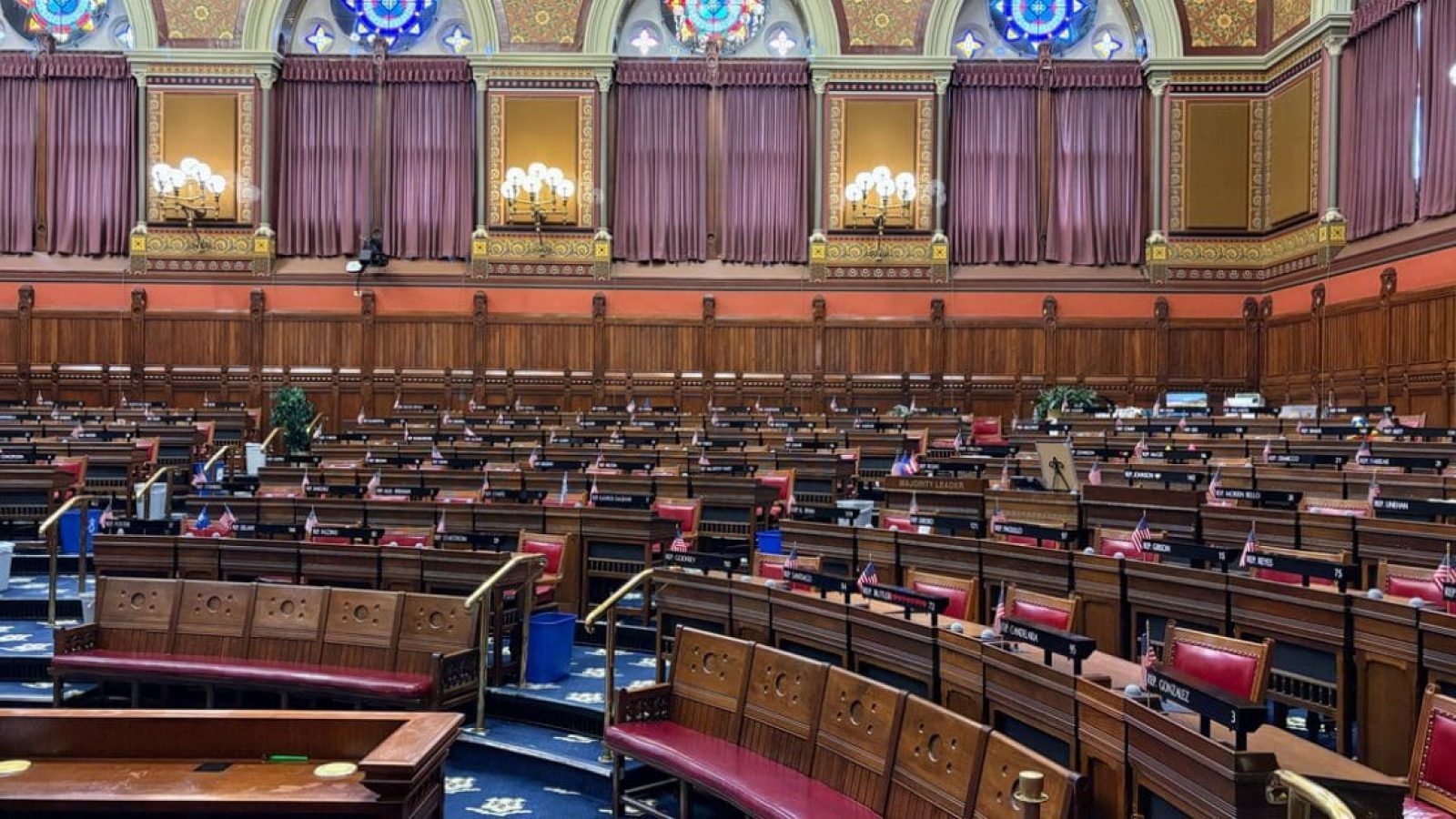 Interior view of a legislative assembly hall featuring wooden seating, decorative stained glass windows, and empty rows of desks with nameplates.
