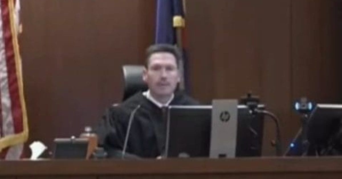 Judge presiding over a court session, seated at a desk with computers and an American flag in the background.
