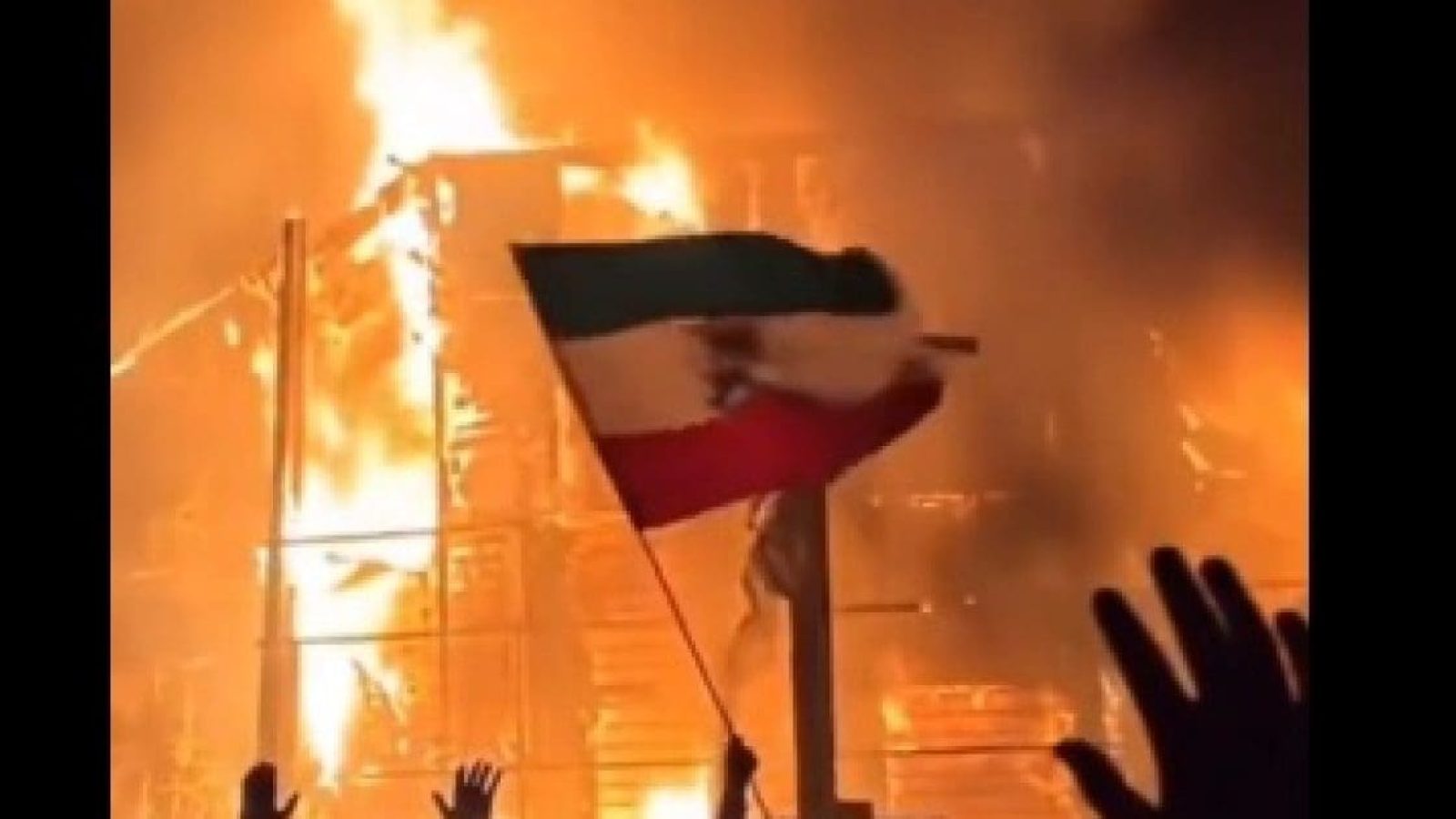 Flames engulf a building as a person waves a Mexican flag, symbolizing protest and unrest during a chaotic event.