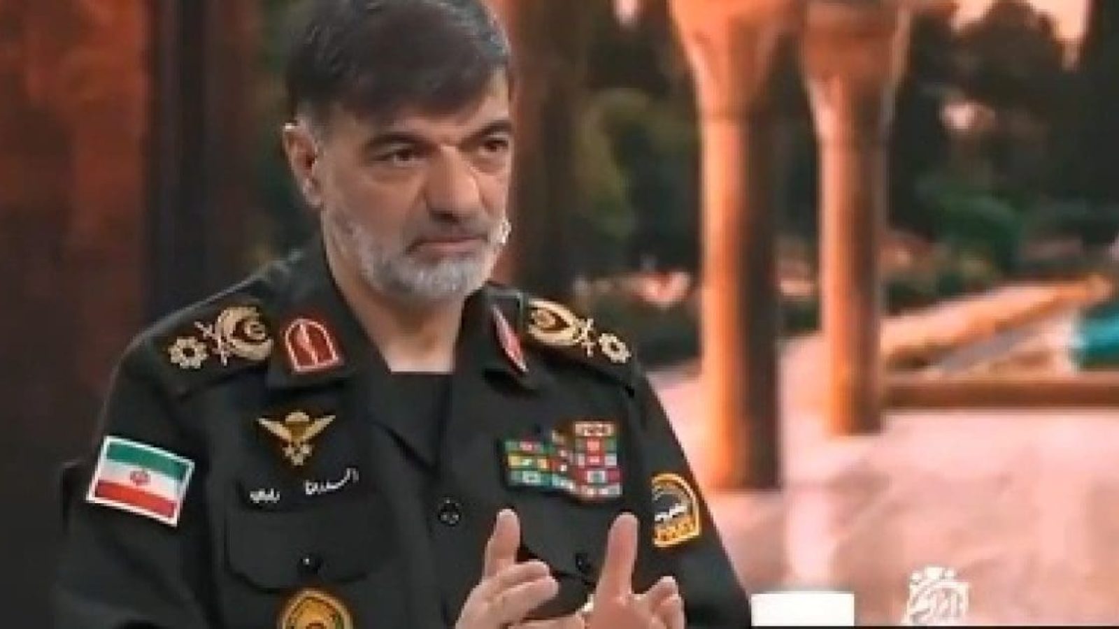 Military official in uniform with medals and Iranian flag, discussing defense topics on a news program set against a scenic backdrop.