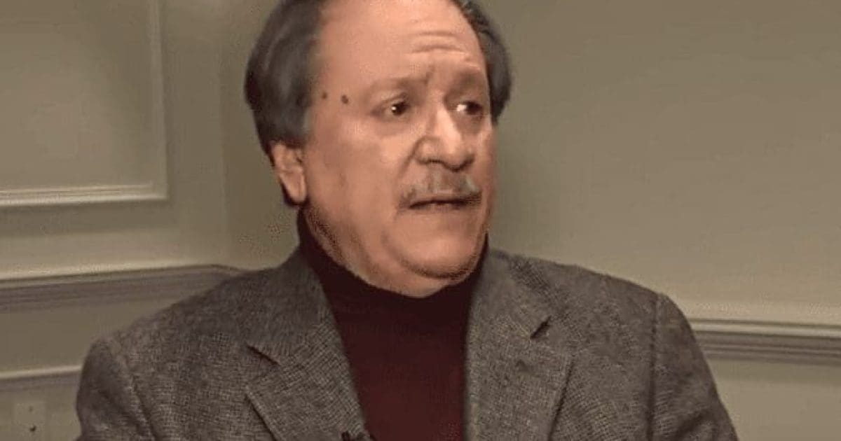 Man in a gray blazer and dark turtleneck speaking during an interview, conveying a serious expression in an indoor setting.
