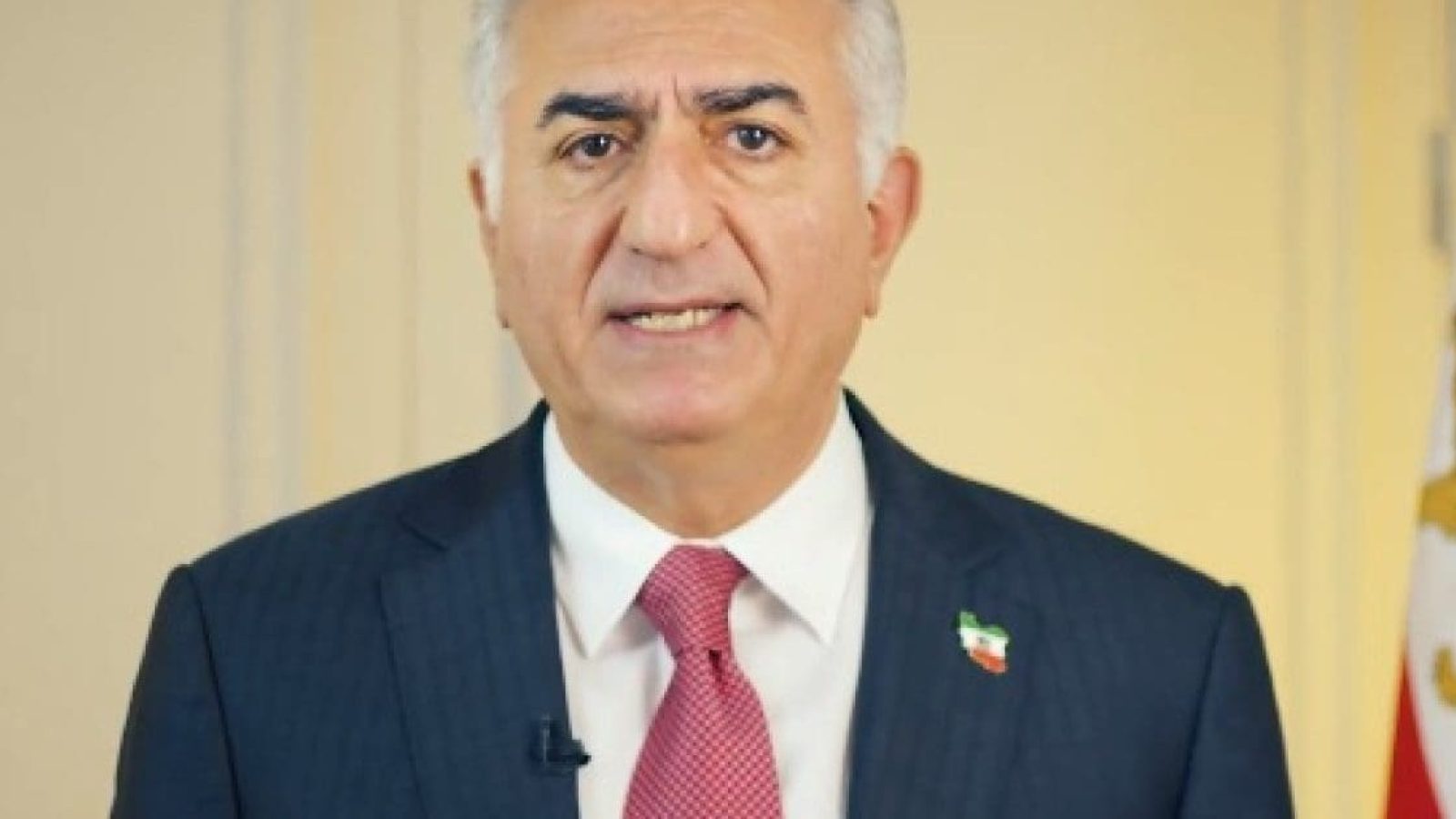 A man in a suit with a red tie addresses the camera, showcasing a flag pin on his lapel, set against a neutral background.