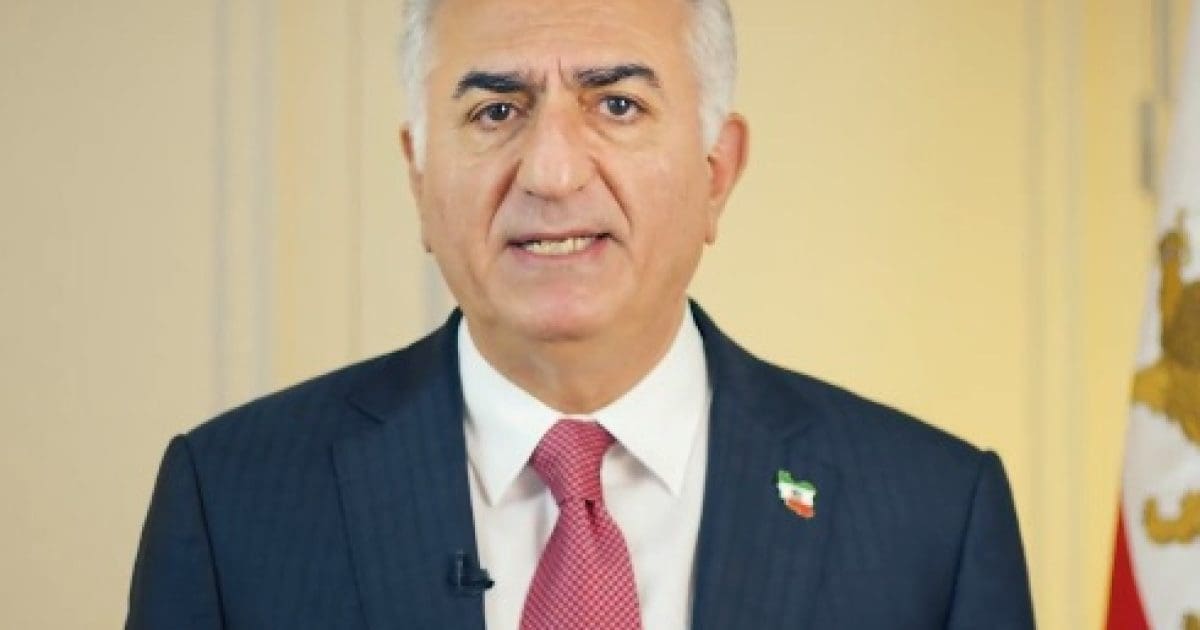 A man in a suit with a red tie addresses the camera, showcasing a flag pin on his lapel, set against a neutral background.