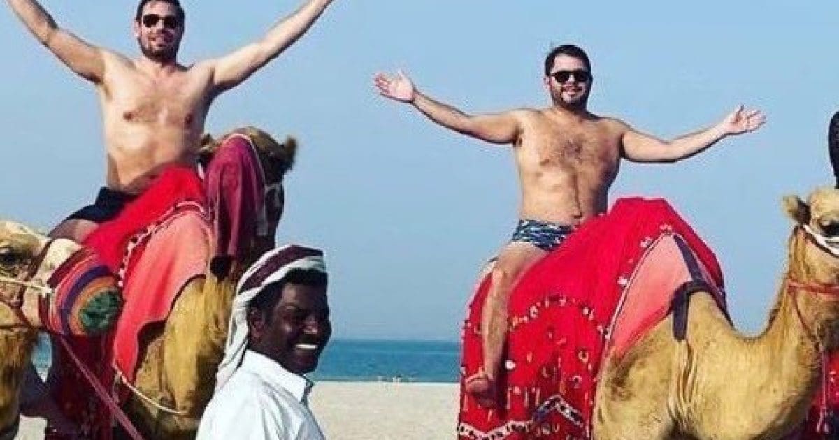 Two men riding camels on a beach, smiling and enjoying the sunny day, with a local guide in traditional attire nearby.
