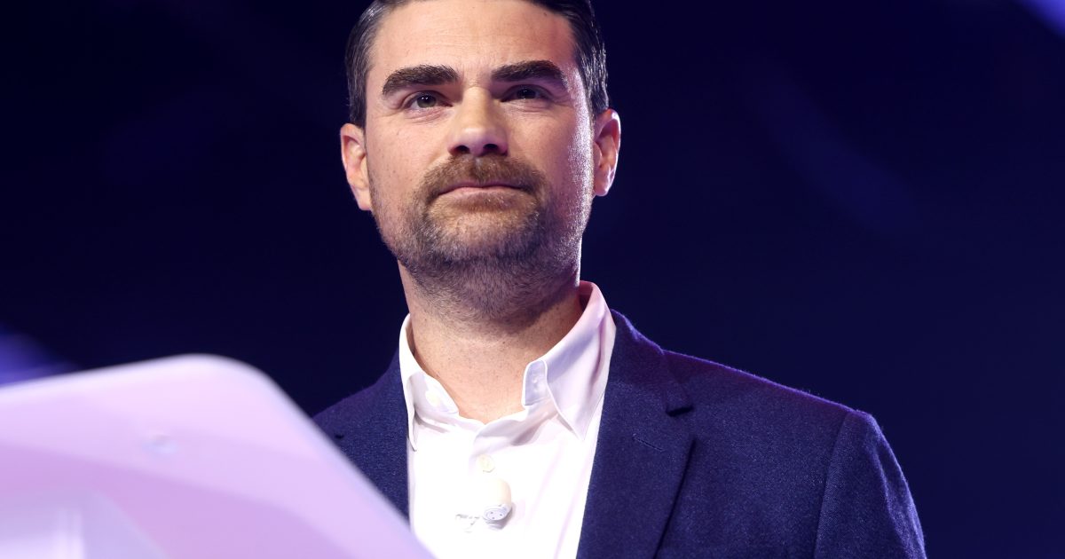 Presenter speaking at an event, wearing a dark blazer and white shirt, with a microphone visible, against a colorful background.