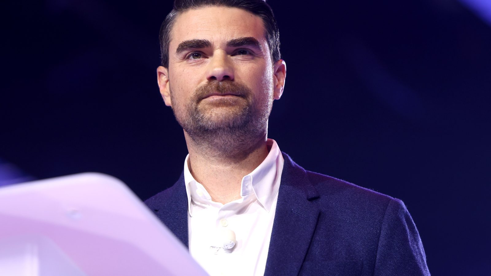 Presenter speaking at an event, wearing a dark blazer and white shirt, with a microphone visible, against a colorful background.