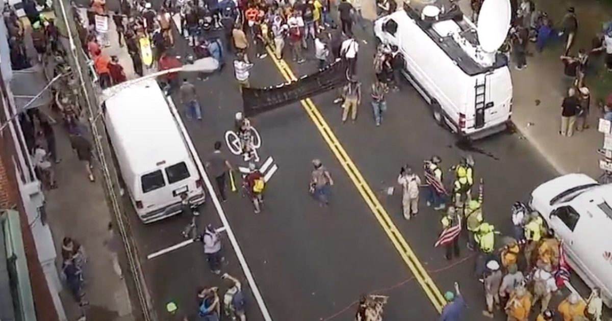 Aerial view of a large crowd gathering in a street, featuring protesters, police vehicles, and media vans during a public demonstration.
