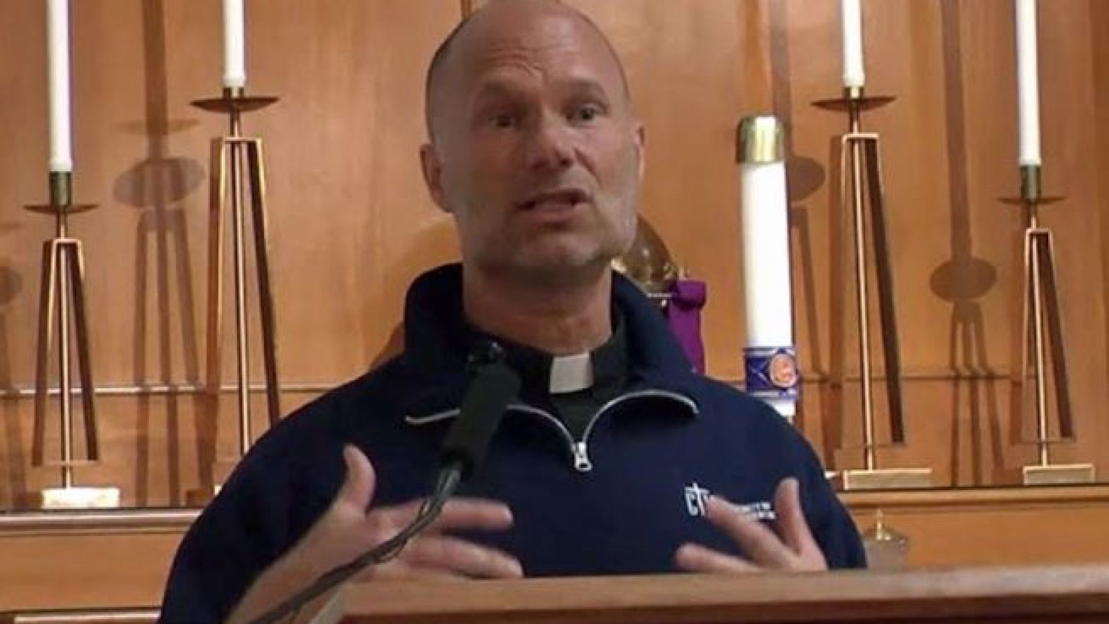 Clergy member delivering a sermon in a church setting, gesturing while speaking to the congregation.