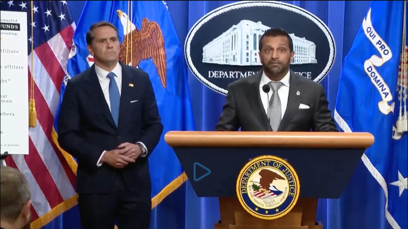 Press conference at the Department of Justice featuring officials discussing financial investigations, with flags in the background and a presentation board visible.