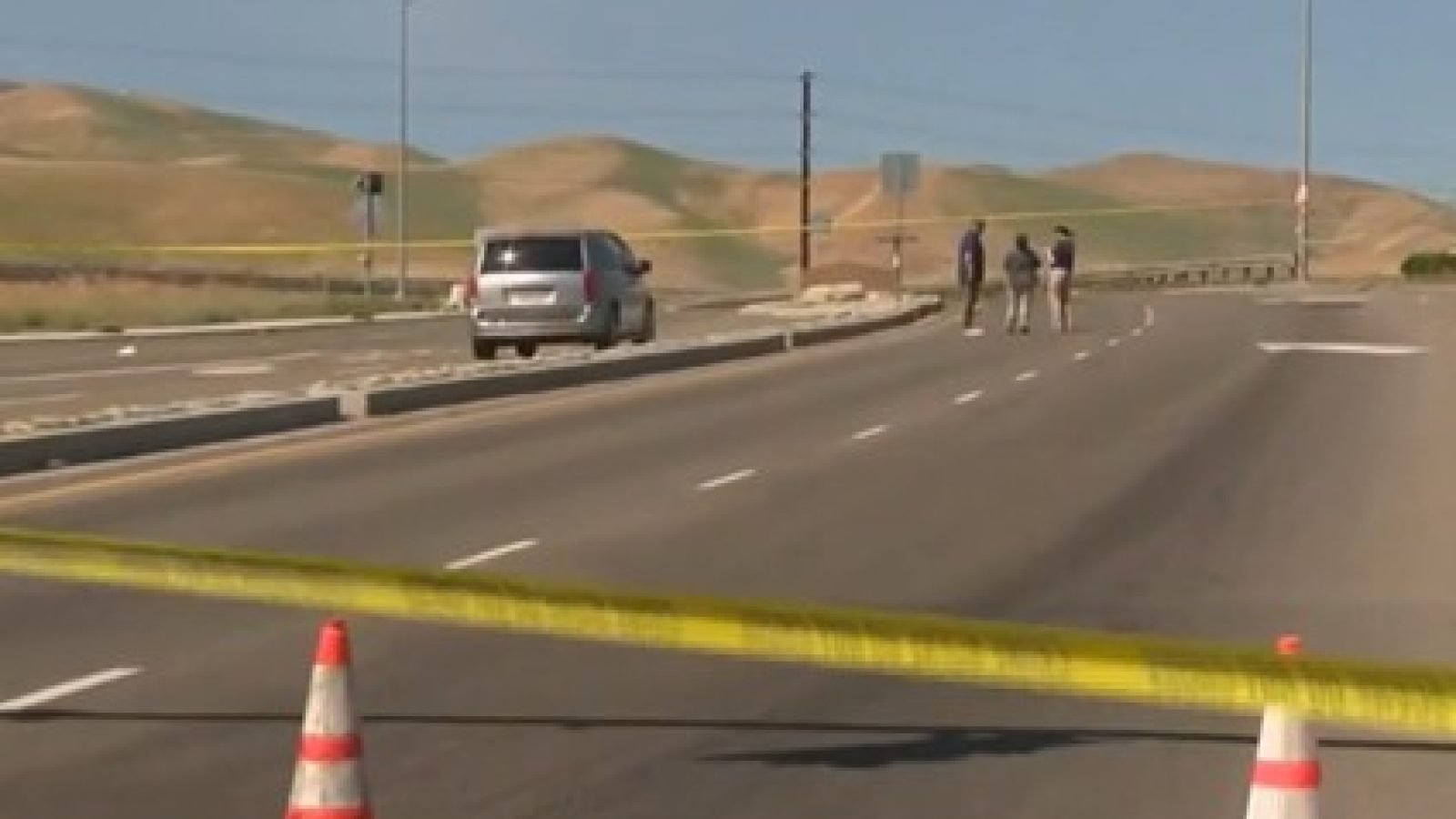 Emergency responders investigate a blocked road scene marked by police tape and traffic cones, with a vehicle parked nearby and hills in the background.