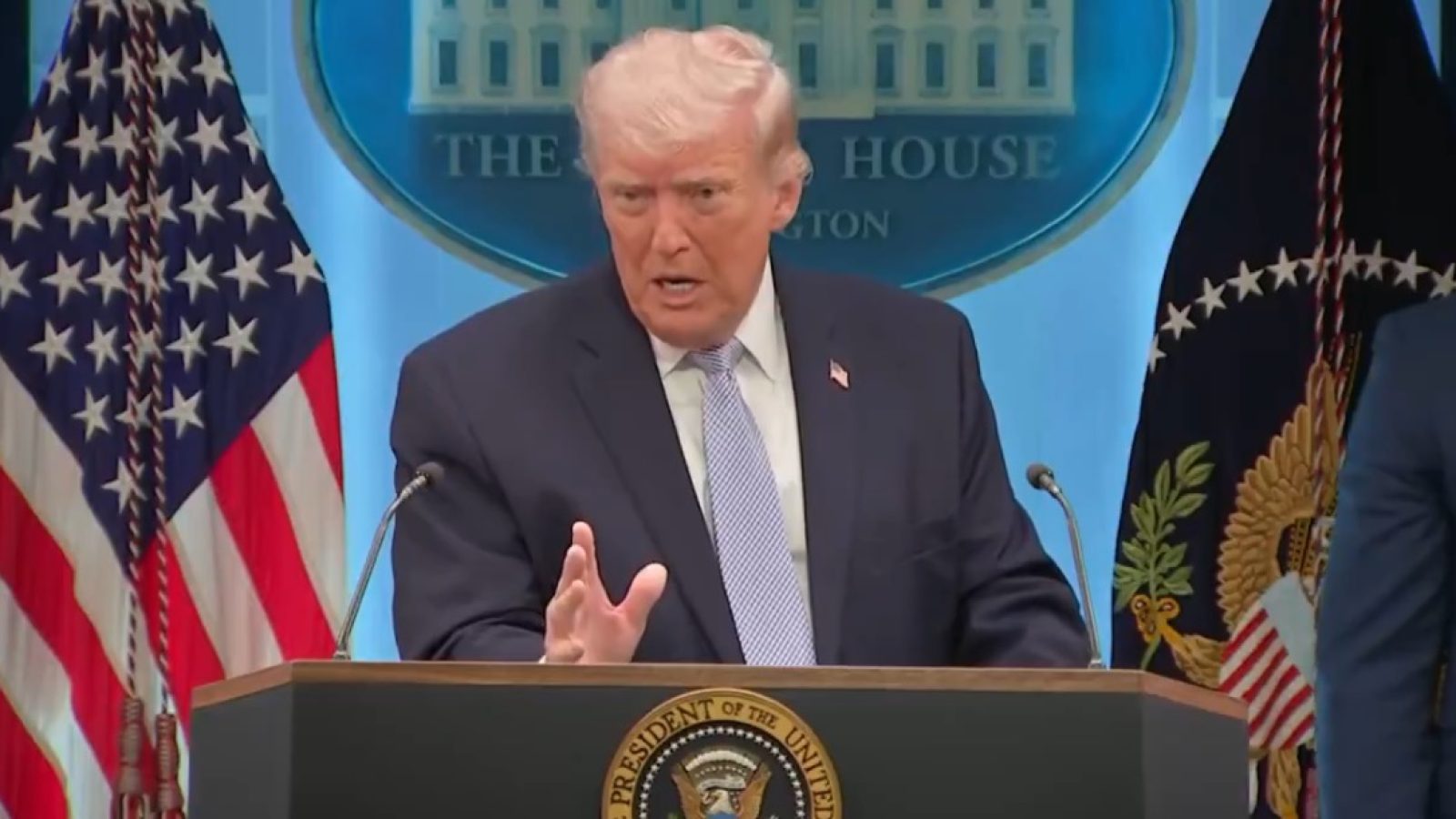 Donald Trump speaks at a press conference in the White House briefing room, flanked by American flags and the presidential seal.