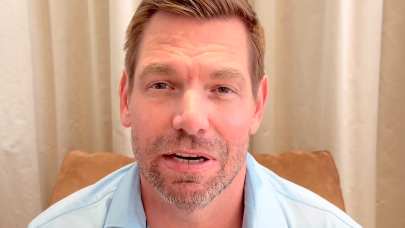 Smiling man in a light blue shirt sitting in front of a beige curtain, speaking directly to the camera in a casual setting.