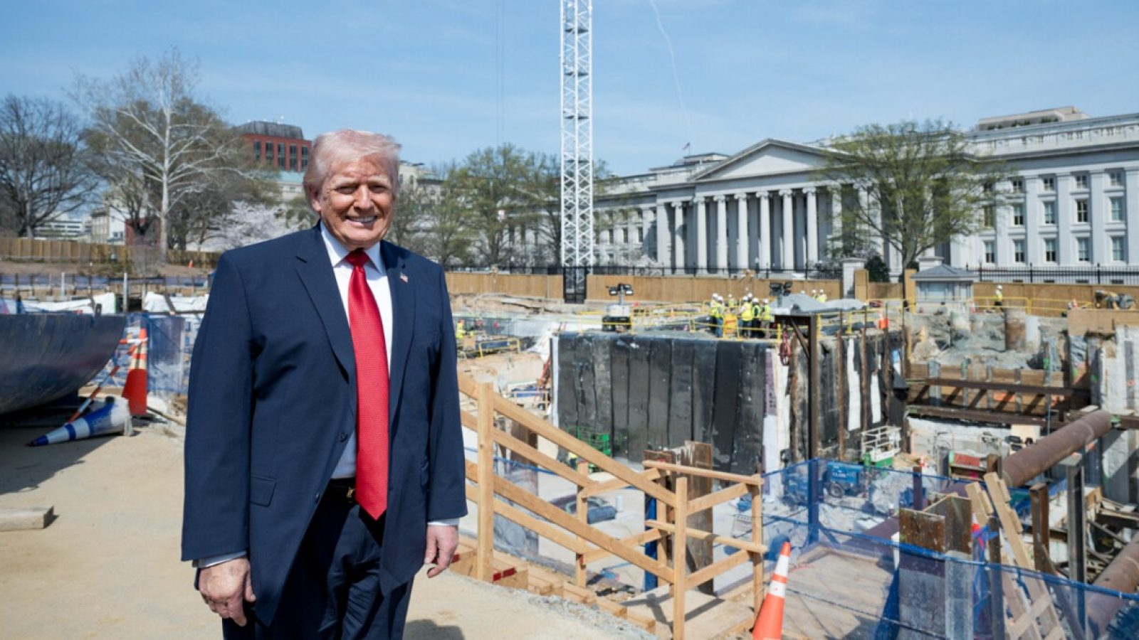 Former President Donald Trump stands in front of construction site near the U.S. Treasury, with workers and equipment visible in the background.