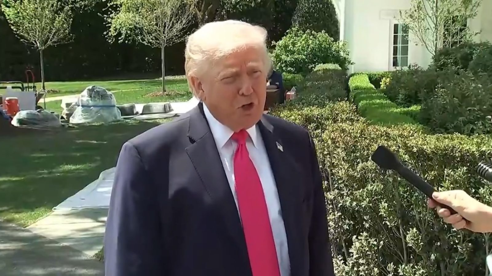 Donald Trump speaking to reporters in the White House garden, wearing a suit and pink tie, with greenery and construction materials in the background.