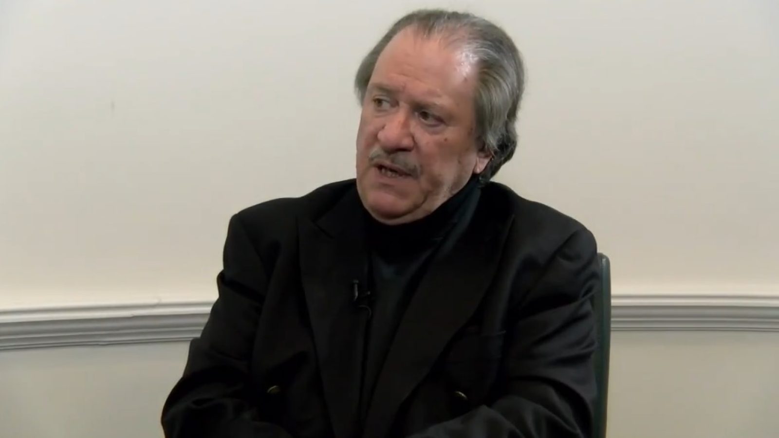 Man in a black jacket sitting in an interview setting, engaged in conversation with a serious expression against a neutral background.