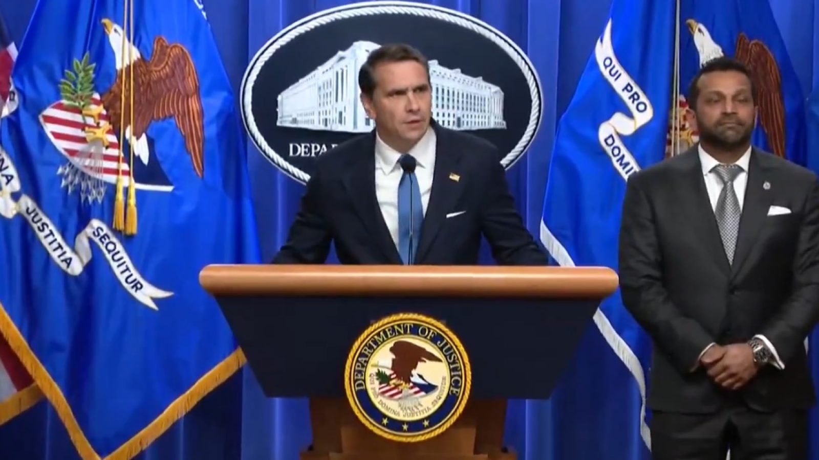 Official announcement by Department of Justice officials at a press conference, featuring a speaker at a podium with the DOJ seal and American flags in the background.