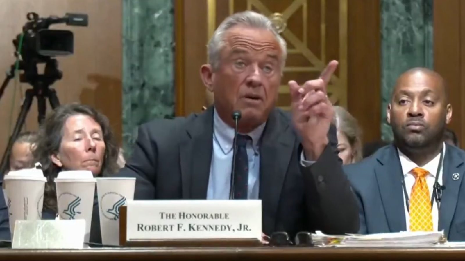 Robert F. Kennedy Jr. speaks passionately during a congressional hearing, with coffee cups in front and attendees listening intently in the background.