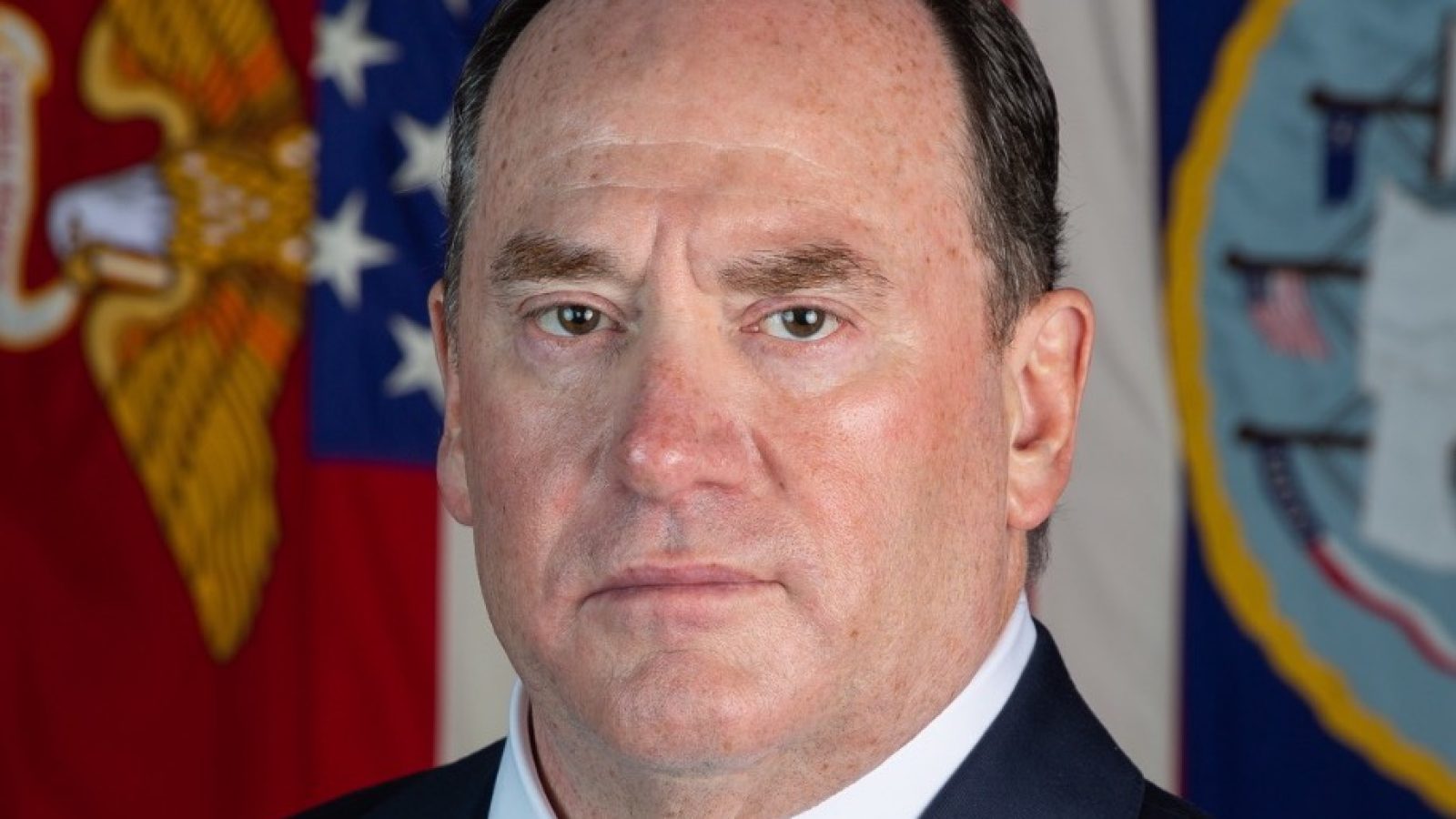 Portrait of a serious-looking man in a suit against a background of military flags, emphasizing authority and professionalism.