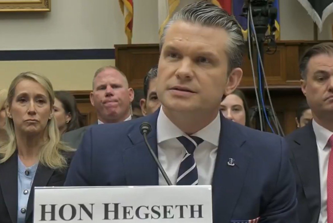 Hearing session with Hon. Hegseth speaking, surrounded by attentive audience members in a formal setting.