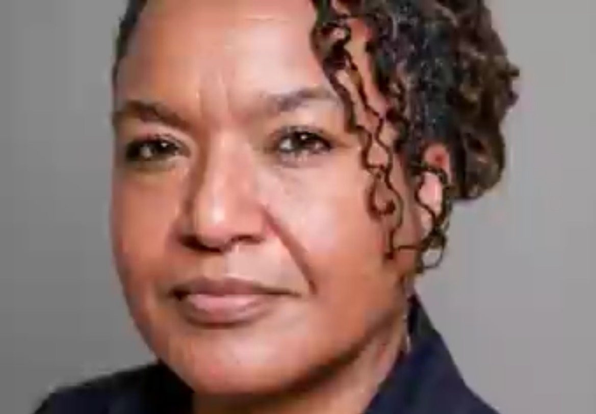 Portrait of a confident woman with curly hair, wearing a dark top, against a neutral background, conveying a professional and approachable demeanor.