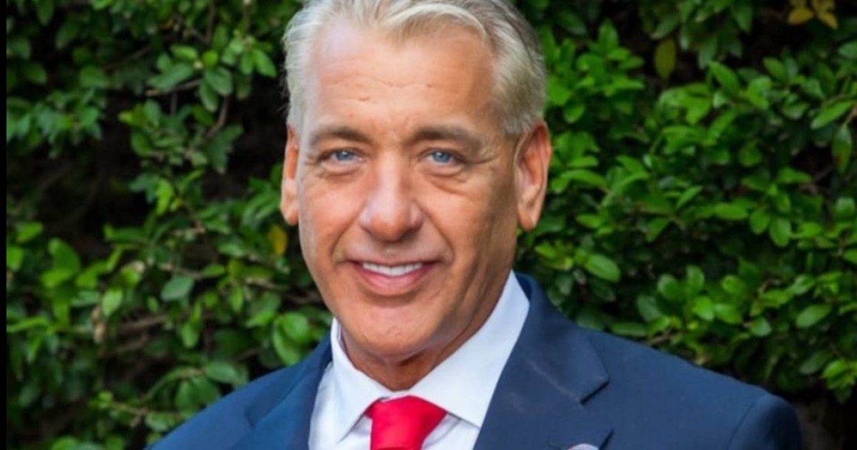 Smiling man in a suit with a red tie stands against a lush green background, exuding confidence and professionalism.