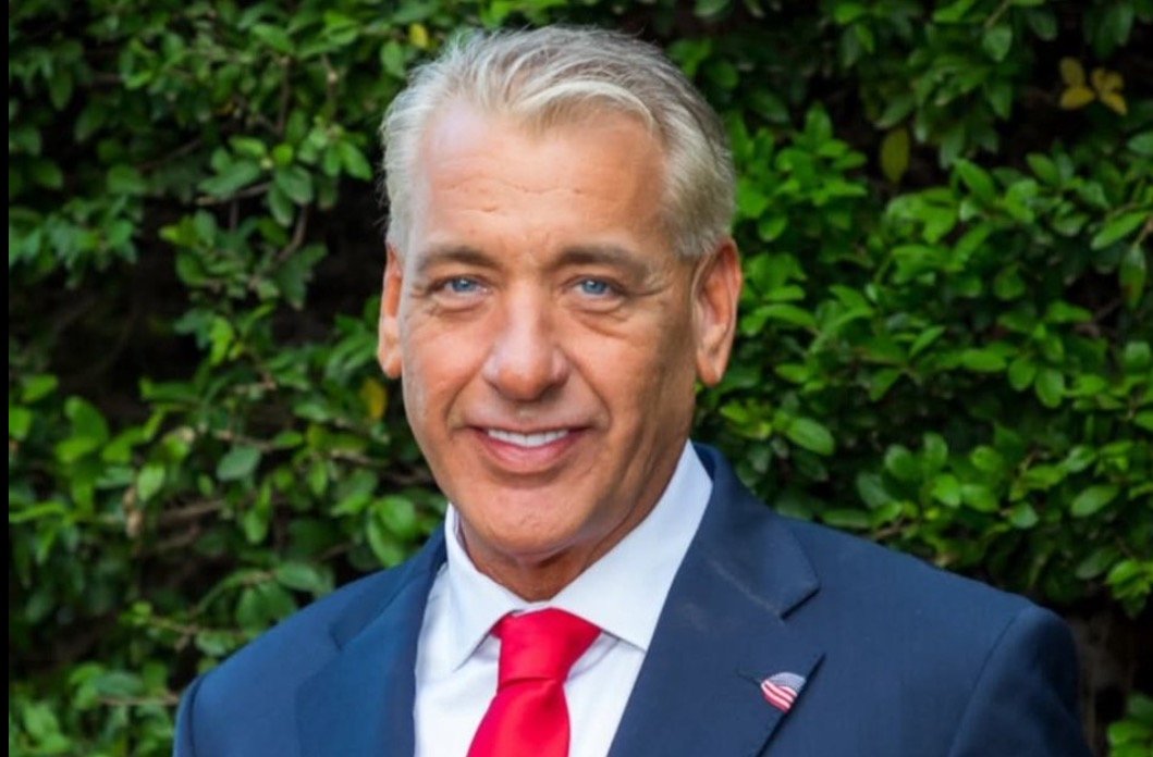 Smiling man in a suit with a red tie stands against a lush green background, exuding confidence and professionalism.