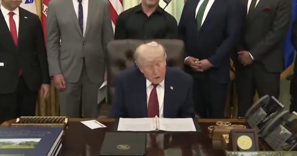 Former President Donald Trump speaking at a desk in the Oval Office, surrounded by advisors and flags, during a formal event.