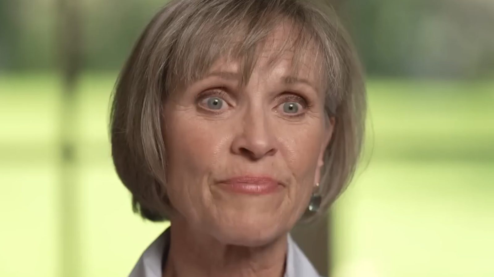 Close-up of a woman with short gray hair and green eyes, expressing a thoughtful emotion in a softly lit indoor setting.