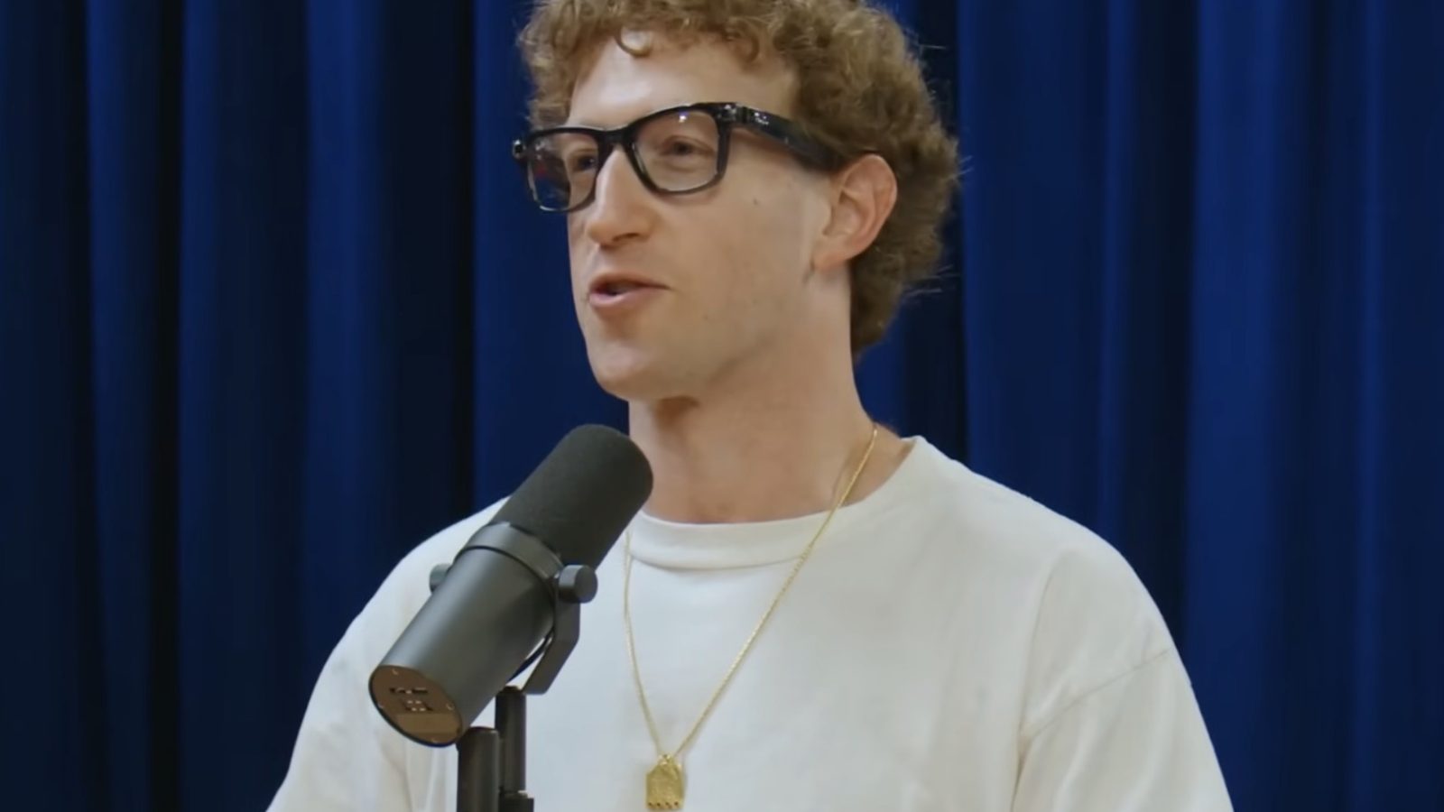 Person speaking at a podcast with a microphone, wearing glasses and a white t-shirt, against a blue backdrop.