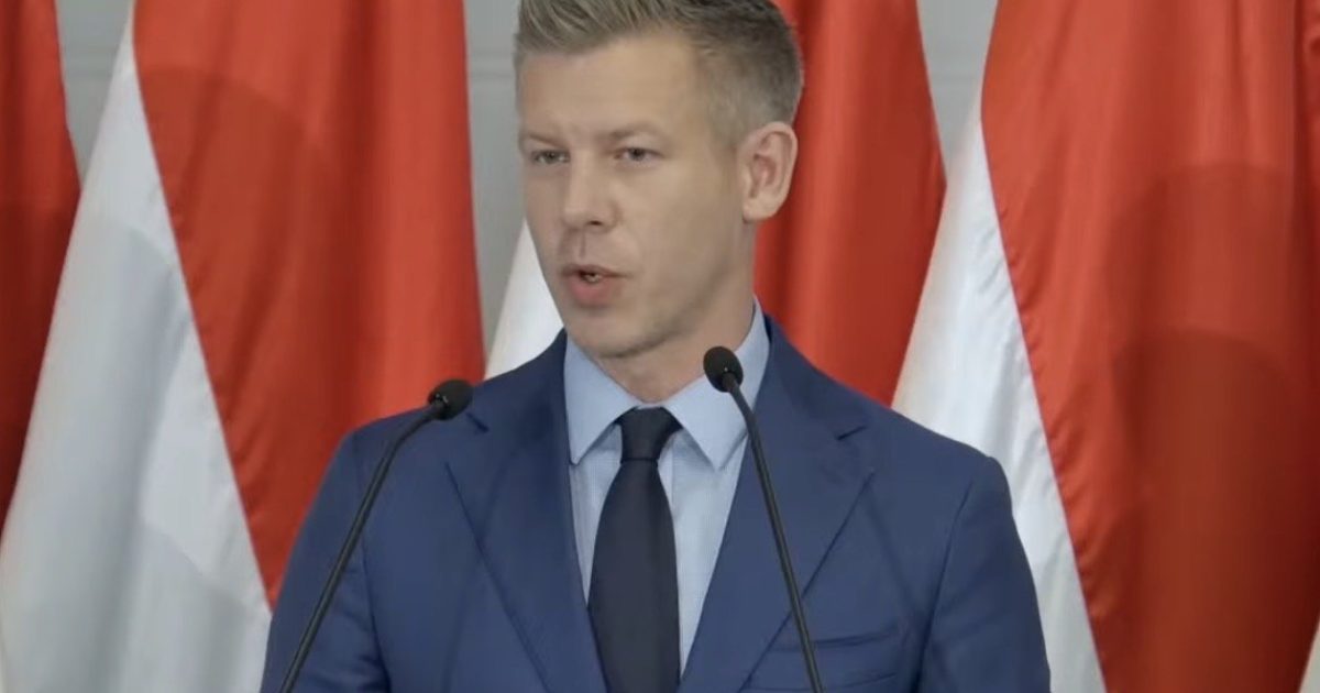 Man in a blue suit speaking at a podium with microphones, surrounded by red and white flags, during a formal event.