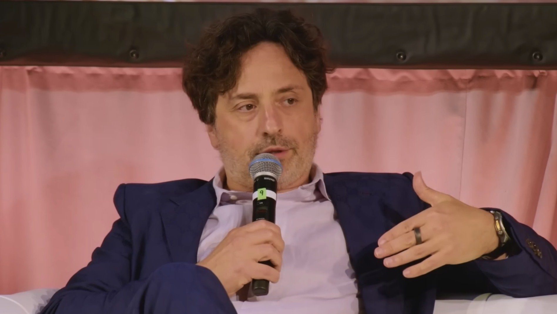 Man in a blue suit speaking into a microphone during a panel discussion, seated on a white couch with a glass of wine nearby.