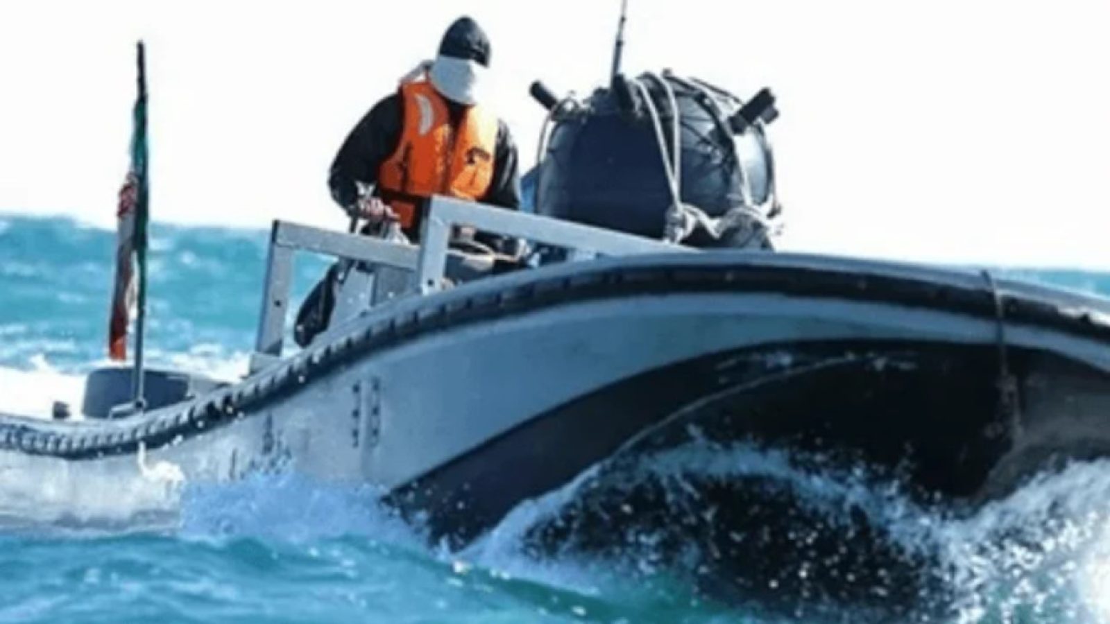 A person in an orange life jacket operates a small boat on choppy blue waters, with a flag visible in the background.