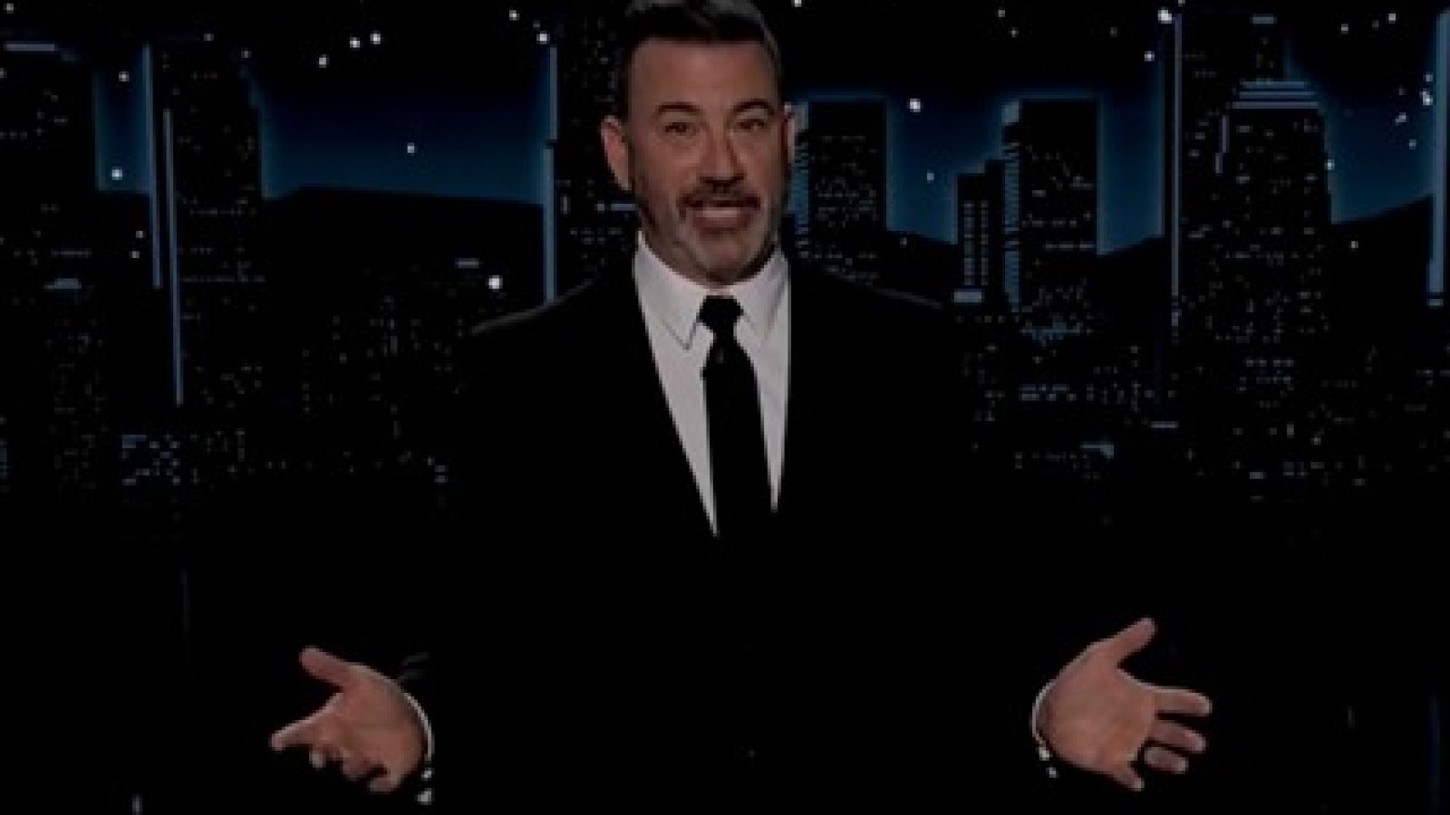 Jimmy Kimmel hosting a late-night show, dressed in a black suit, gesturing with his hands against a city skyline backdrop.