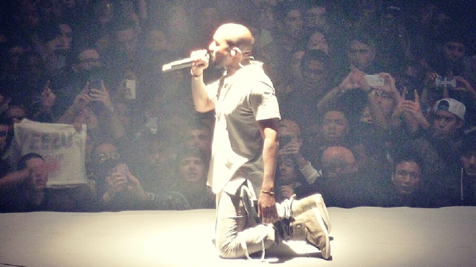 A performer kneels on stage with a microphone, surrounded by an enthusiastic crowd capturing the moment on their phones.