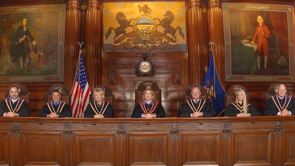 Group of judges seated in a courtroom, with American and state flags, surrounded by historical paintings.