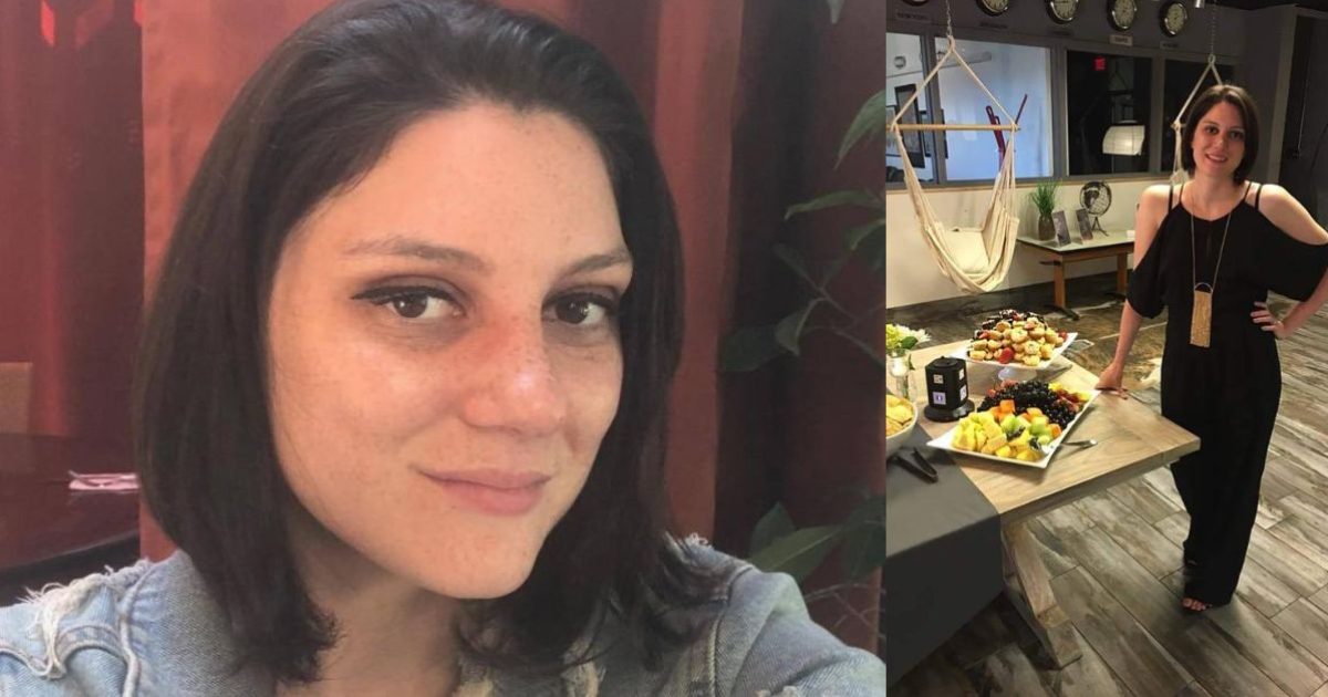 Woman with shoulder-length dark hair smiles at the camera, while standing beside a table filled with a variety of colorful fruits and snacks.
