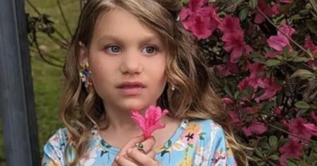 Young girl holding a pink flower, standing in front of vibrant pink azalea bushes, wearing a floral dress and looking thoughtfully into the distance.